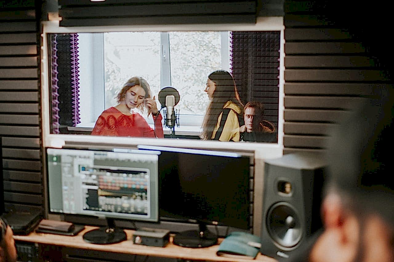Artists recording music in a professional studio setting, captured through a glass window.