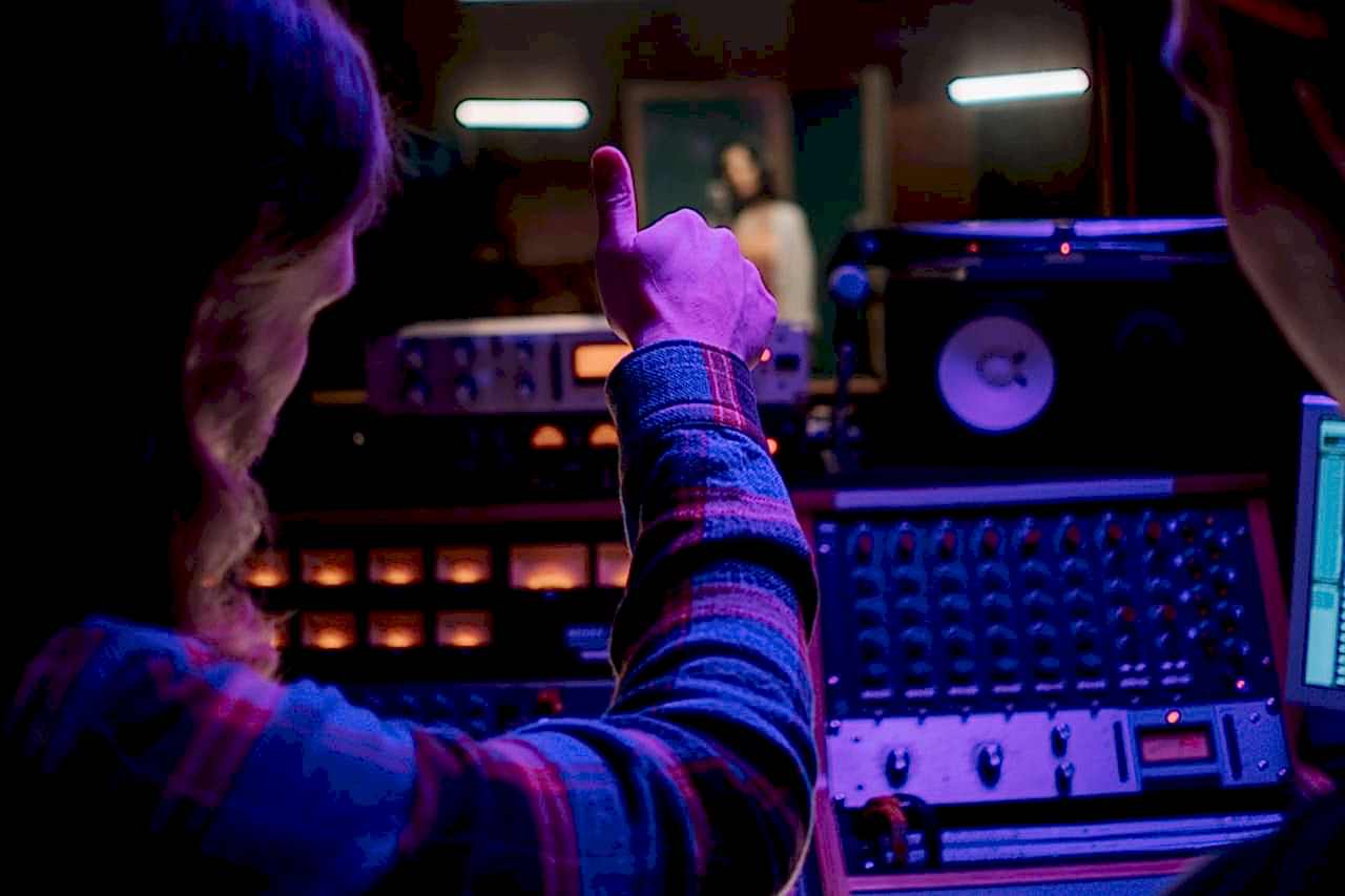 Audio engineer gesturing thumbs up in a vibrant music studio with equipment.