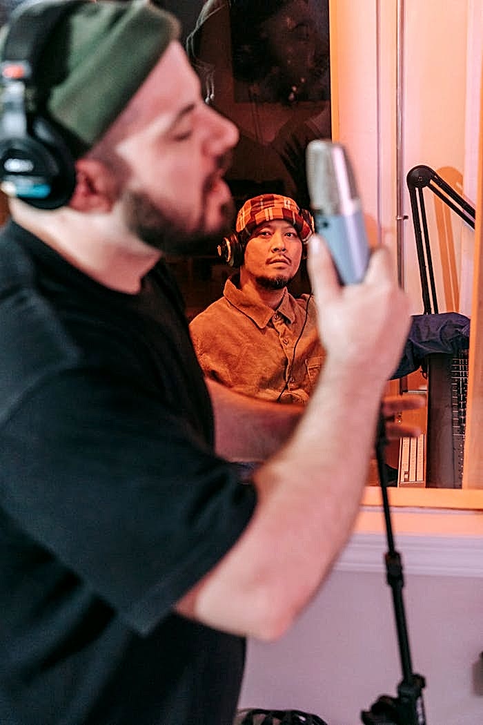 Two men collaborate in a recording studio. One sings into a microphone while the other observes.